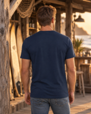 Back view of a man wearing a plain navy Bella+Canvas 3001 T-shirt beside a wooden surf shack.