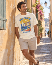 Man leaning against a pastel wall in a Saint-Tropez side street wearing the ivory Saint-Tropez Côte d'Azur tee