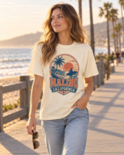 Woman wearing the ivory Malibu California Surf Tee while walking along a beachfront promenade at sunset