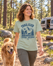 Woman wearing the Bay Trail Club tee with a navy hiker and dog graphic, walking beside a dog at a pine forest campsite.