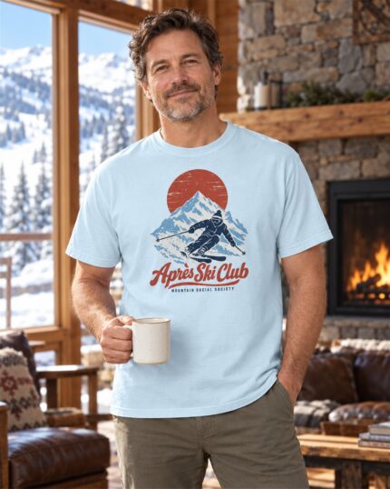 Man wearing the Chambray Après Ski Club Skier T-shirt by a lodge fireplace with a warm mug