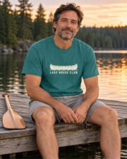 Man in the Emerald Lake House Club Canoe T-shirt sitting on a lake dock at sunset