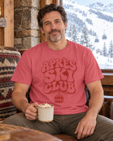 Man wearing the Watermelon Après Ski Club Retro T-shirt by a lodge window with a hot chocolate
